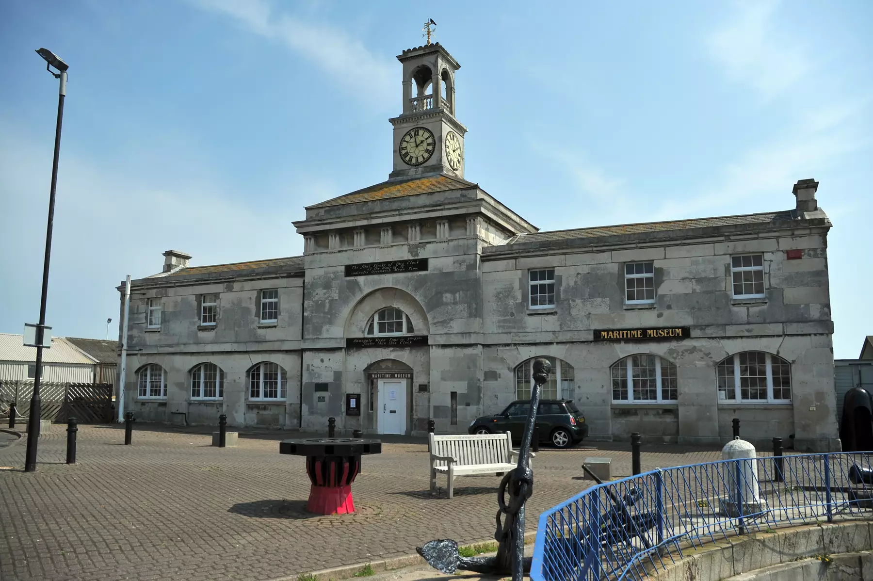 Exterior of Ramsgate Maritime Museum built of grey blocks with white windows on both floors and a white front door. Clock tower with giant beige clock face and black arms and roman numerals. Above this bell tower with a gold weather vane.